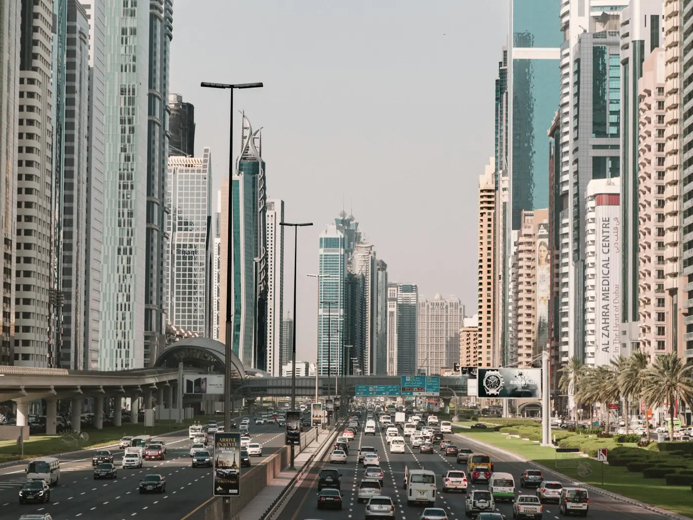 Traffic on a multi-lane road between skyscrapers in Dubai, with the Dubai Metro line alongside